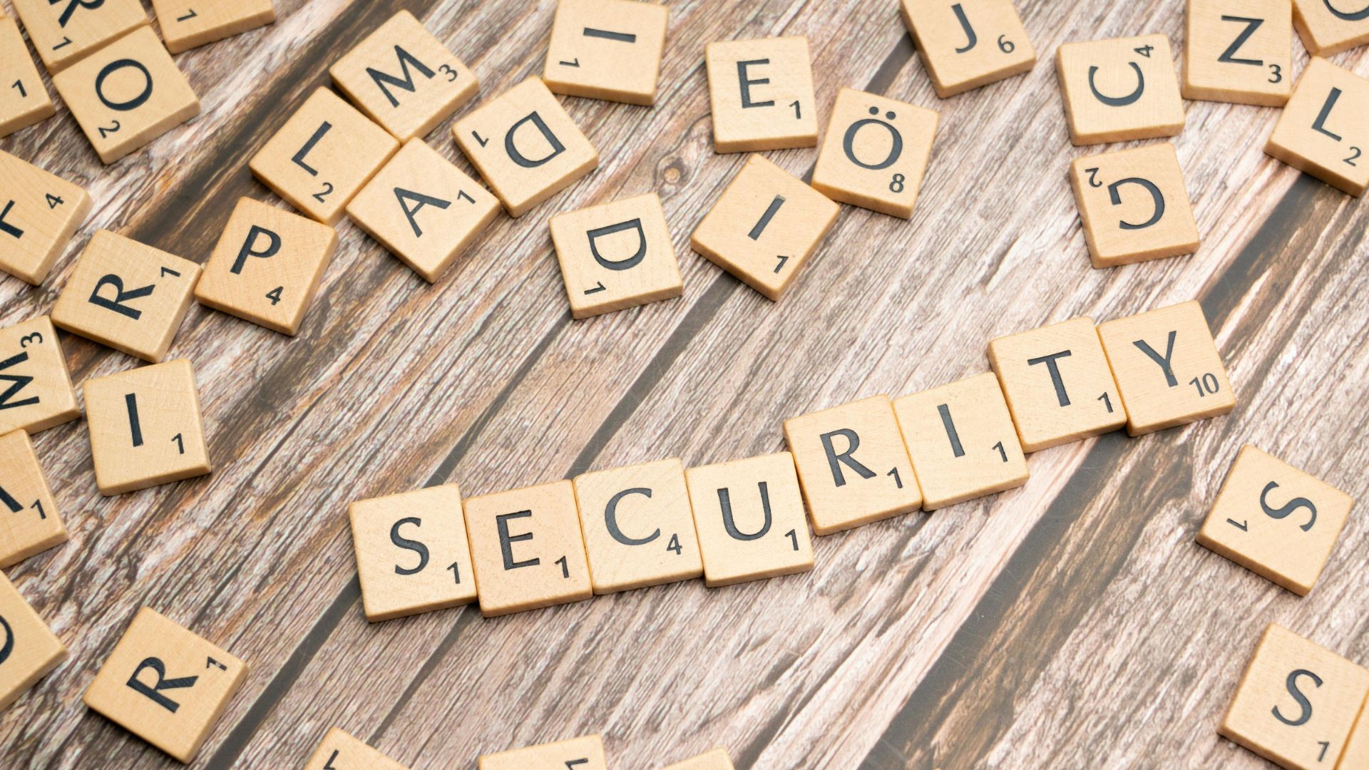 Wooden Scrabble tiles arranged to spell “SECURITY” on a wooden surface, surrounded by other scattered tiles.