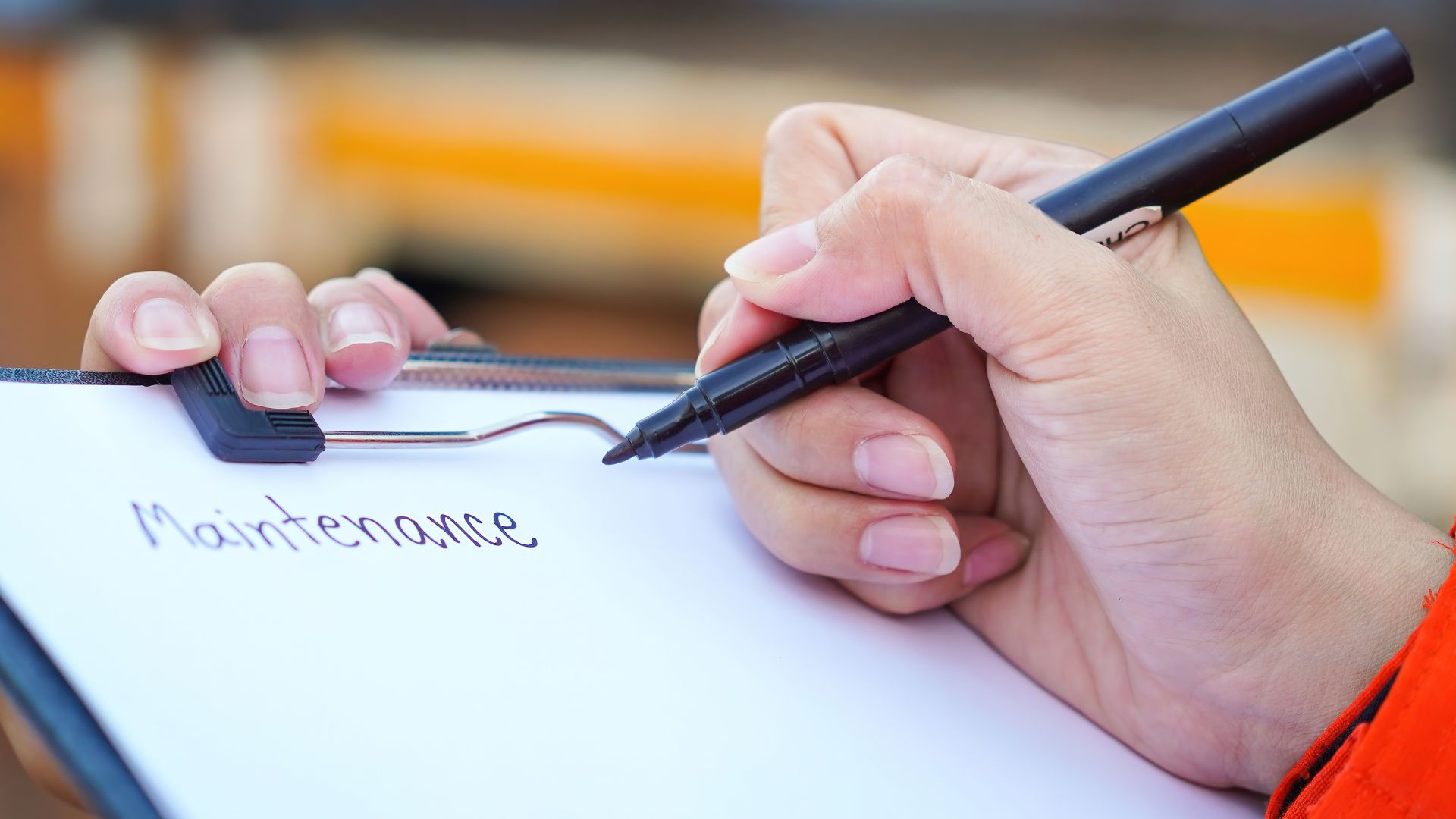 Close-up of a person's hand writing “Maintenance” on a clipboard with a marker, indicating scheduled checkups or upkeep tasks.