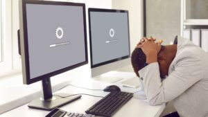 Frustrated person with head in hands at a desk while computers display slow loading bars.|Hand placing a wooden block labeled “Plugin” between others